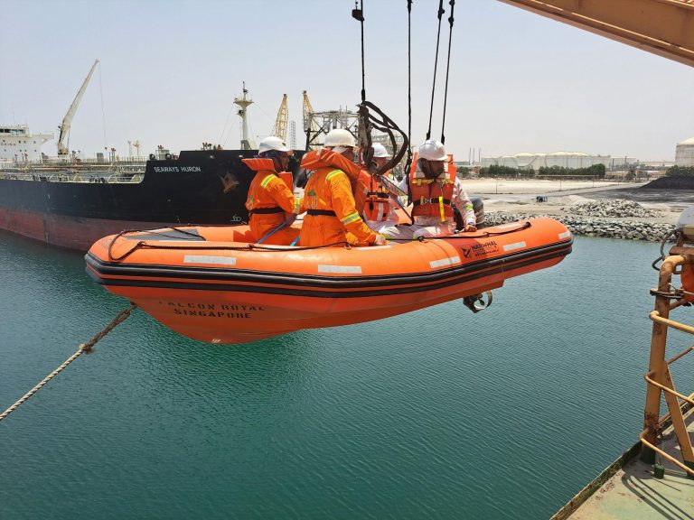 Crew members in safety gear on a lifeboat being lowered at a harbor in Sharjah, UAE.